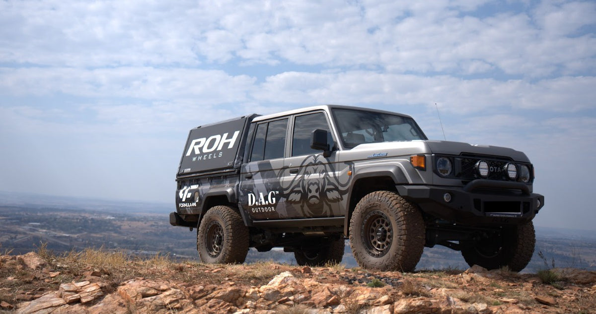 A black off-road pickup truck with custom graphics and decals, including 'ROH WHEELS' and 'D&G CUSTOMS', parked on a rocky hillside under a cloudy sky.