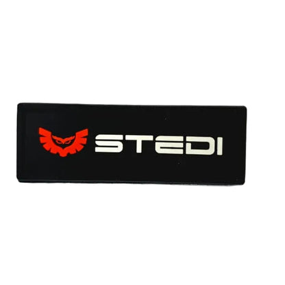 A black rectangular patch featuring the word 'STEDI' in white alongside a red devil-like logo, placed on a white graph paper background.