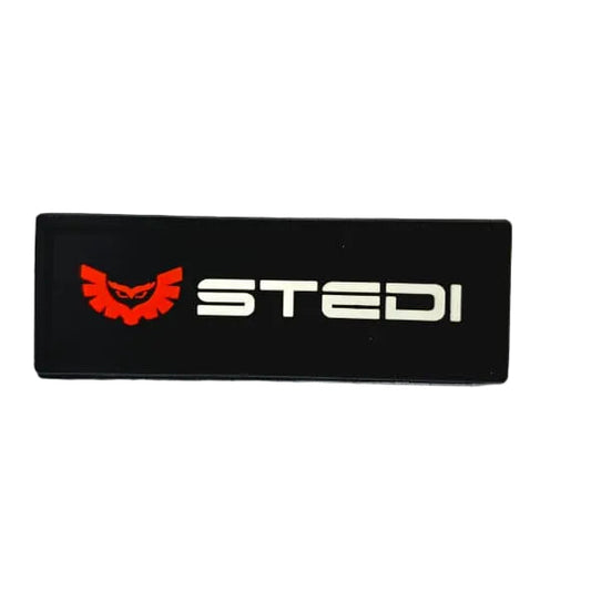 A black rectangular patch featuring the word 'STEDI' in white alongside a red devil-like logo, placed on a white graph paper background.